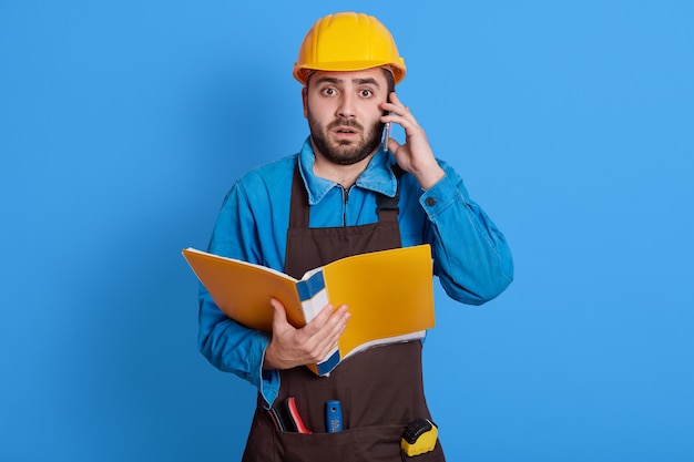 Workers wearing safety helmet, apron and uniform talking on mobile phone, having shocked facial expression, has problems on building, needs solving troubles, holding paper folder in hands.