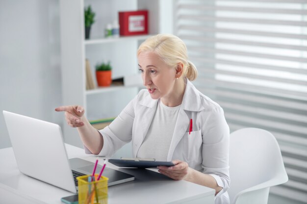 Workflow, medic. Serious woman in medical coat holding a folder, pointing with her hand away, looking at the laptop screen.