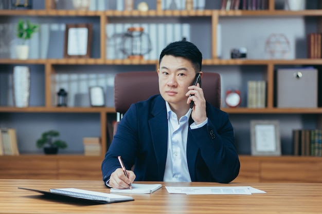 ワークフロー若いハンサムな成功したアジア人男性が電話で話している電話を使用してオフィスで働いている文書を扱うテーブルに座っている銀行家の弁護士