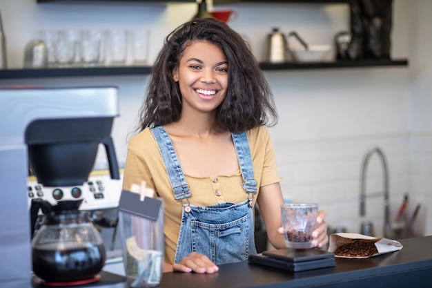 営業日、カフェ。コーヒーショップのカウンターの後ろに立っているガラスのコーヒー豆と幸せな若いムラートの女性