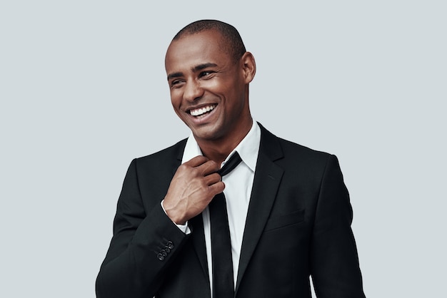 Working day is over. Charming young African man in formalwear adjusting necktie and smiling while standing against grey background