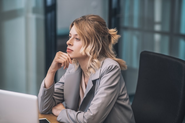 就業日。オフィスの職場に座っているビジネススーツの若い大人の物思いにふける女性