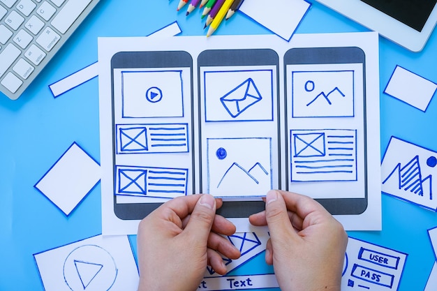 Working desk with hands sorting wireframe screens of mobile responsive website Developing wireframe sketch layout design mockup on smartphonetablet screen