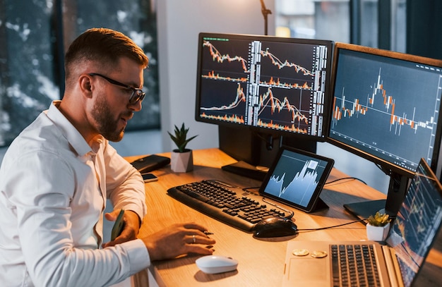 Working at evening time young businessman in formal clothes is\
in office with multiple screens