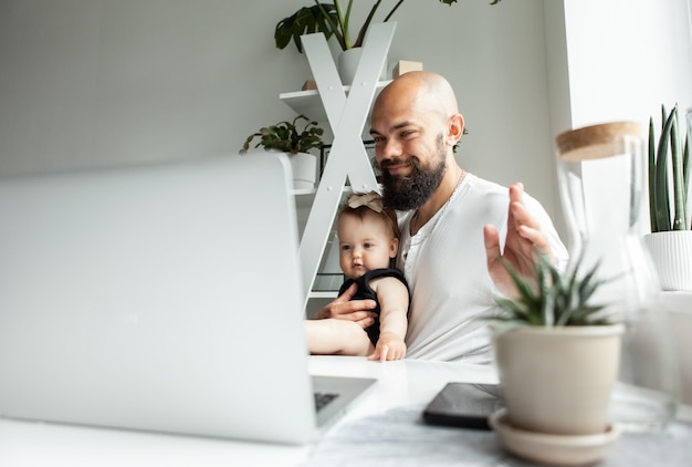 Working father waves to the laptop camera with his little daughter in his arms in a modern room