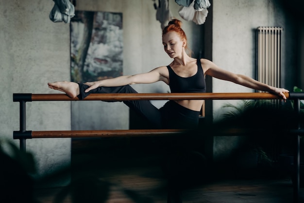 Photo working on flexibility. young red haired woman ballerina lifted one leg on ballet barre and doing stretching exercises against darken background of studio or classroom. barre workout concept