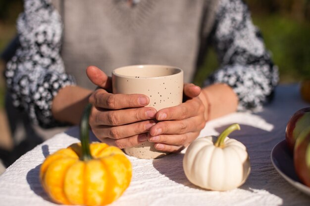 Photo the working hands of the girl hold a ceramic cup standing on the table near small pumpkins