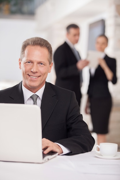 Working on laptop. Cheerful mature man in formalwear sitting at the table and using computer while two colleagues discussing something on background