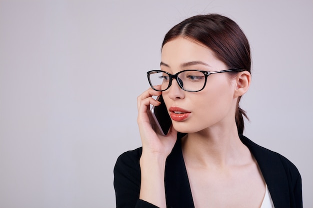 Working a lot. Busy worker. Copy space. Pleasant-looking calm woman with cell phone in her right hand is standing on a gray back in a black jacket, T-shirt and glasses.