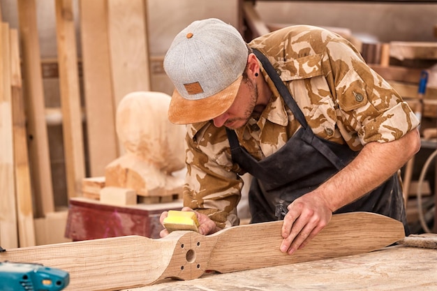 帽子とシャツを着た働く男性が、バックグラウンドツールとボール盤のワークショップで絵を描く前に、サンドペーパーで木製のブロックを磨きます