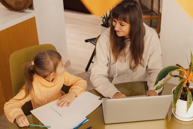 Working mom works from home office with kid woman using laptop child drawing woman freelancer