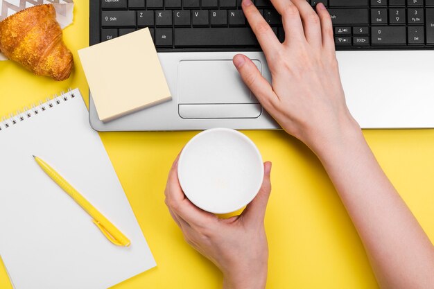 Working morning. Girl works for laptop. Breakfast at work - cappuccino, croissant, notebook on yellow background. Top view