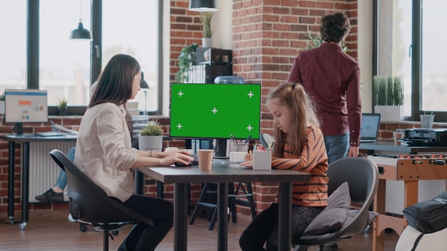 コンピューターで水平方向の緑色の画面を使用している働く母親と机で落書きをしている少女。オフィスで子供を持っている間、孤立したモックアップテンプレートでクロマキーを見ているビジネス女性。