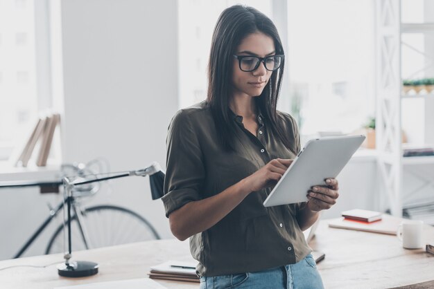 Working in office. Confident young woman in smart casual wear working on digital tablet while standing near her working place