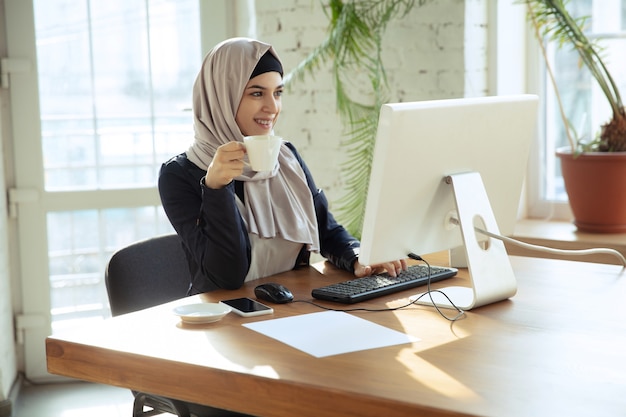 Working online. Portrait of a beautiful arabian businesswoman wearing hijab while working at openspace or office. Concept of occupation, freedom in business area, leadership, success, modern solution.