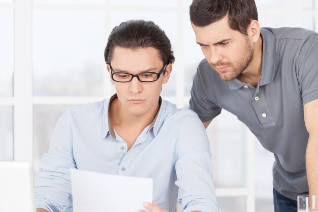 Working out problems. confident young man sitting at the table and looking at the document while his colleague standing near him