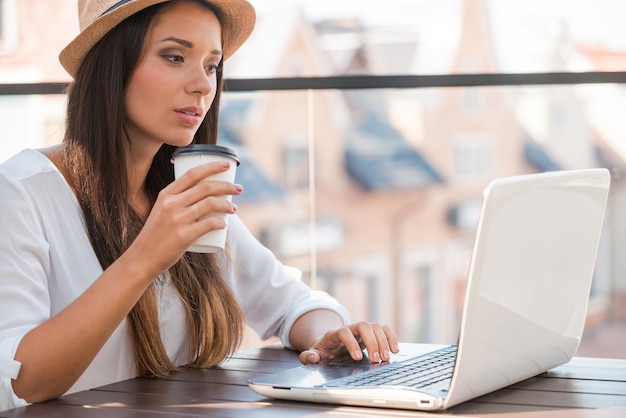 Lavorare all'aperto. bella giovane donna con cappello funky che lavora al computer portatile e sorride mentre è seduta all'aperto