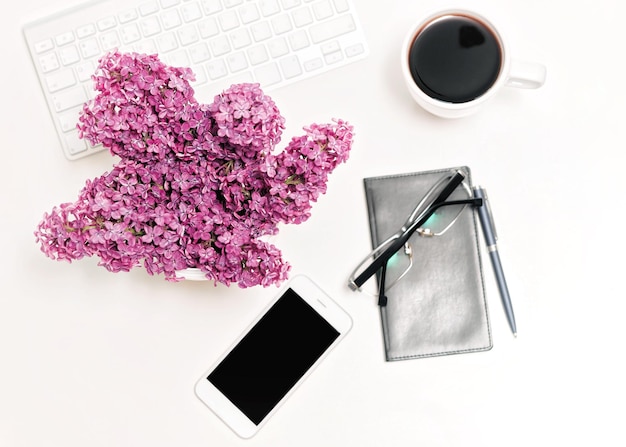 Photo working place with computer keyboard, mobile phone, notebook, cup of coffee, glasses, pen and lilac flowers on the white table background. flat lay, top view