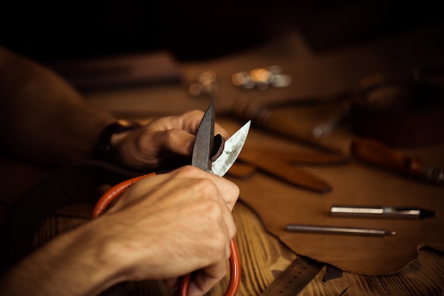 Working process of the leather belt in the leather workshop. Man holding crafting tool and working. Tanner in old tannery. Wooden table background. Close up man arm