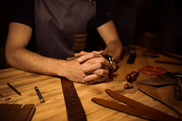 Working process of the leather belt in the leather workshop. Man holding hands on wooden table. Crafting tools on background. Tanner in old tannery. Close up men arm.