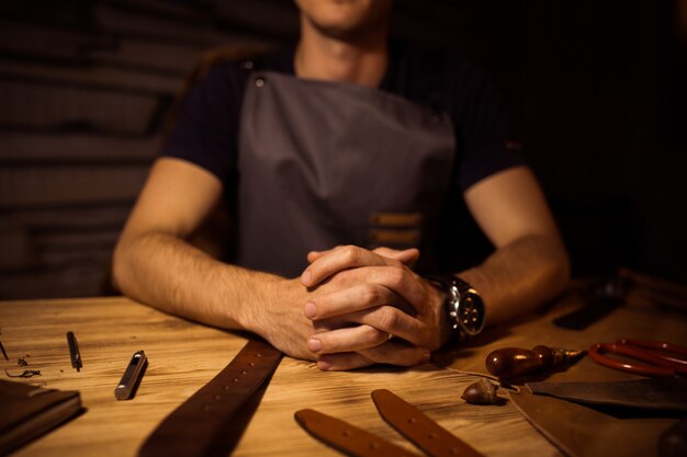 Working process of the leather belt in the leather workshop. Man holding hands on wooden table. Crafting tools on background. Tanner in old tannery. Close up men arm.