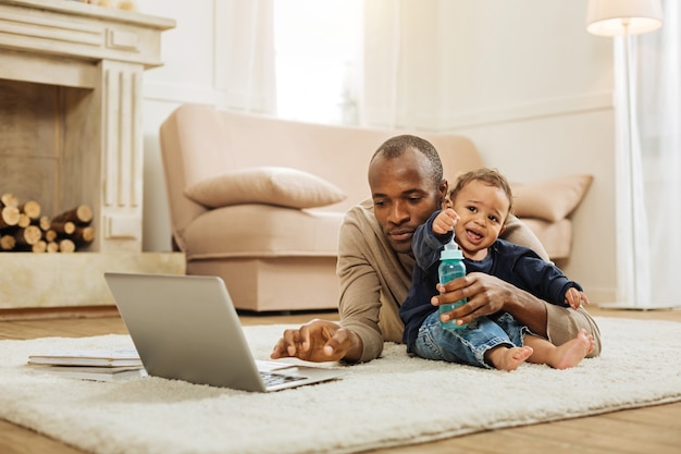 Working on project. Attractive concentrated dark-haired afro-american man working on his laptop on the floor and hugging his little son and holding a bottle