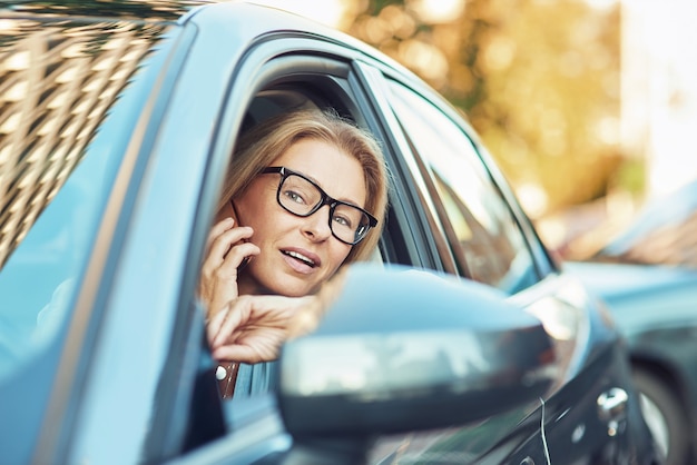 Работая за рулем, зрелая деловая женщина в очках разговаривает по мобильному телефону во время вождения