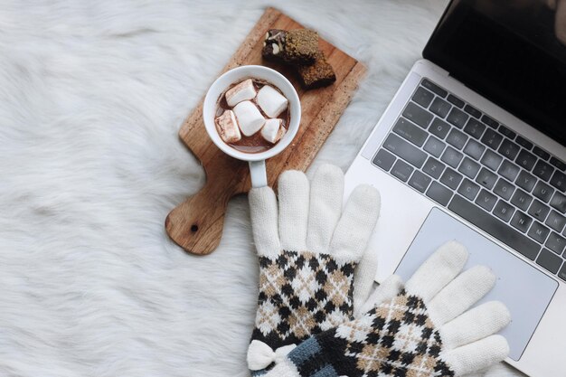 Working in winter. Laptop, mittens and a cup of hot chocolate drink with brownie on white furry rug.