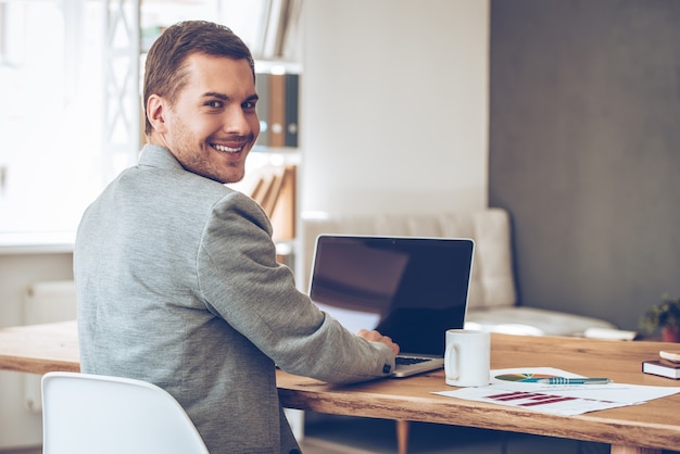 Working with joy. Rear view of cheerful young handsome man looking over shoulder at camera with smile while sitting at his working place