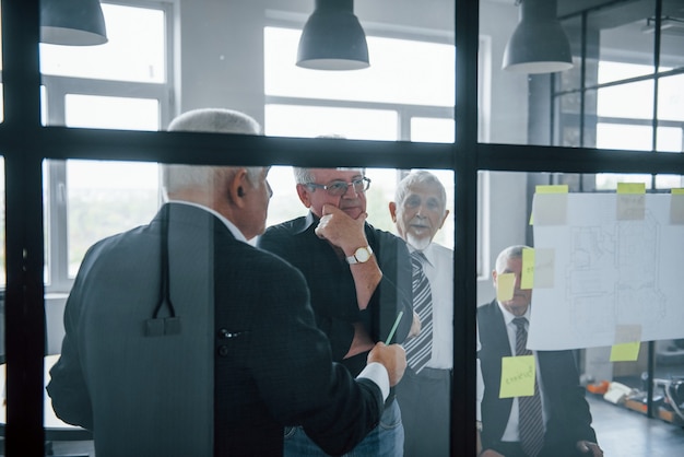 Working with stickers on glass. Aged team of elderly businessman architects have a meeting in the office.