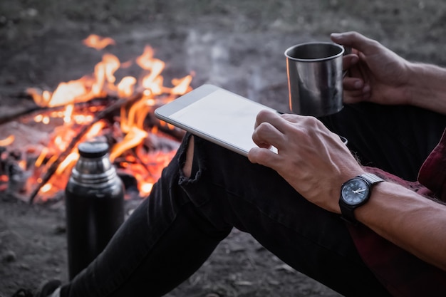 Working with technology in unconventional place concept. Using a tablet computer to work remotely at a hiking trip next to camping fire.