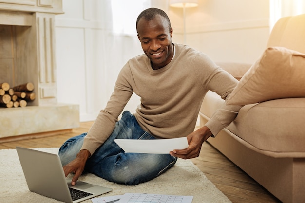 Workload. Happy bearded afro-american man smiling and working on the laptop and holding a sheet of paper while sitting on the floor and a fireplace in the background