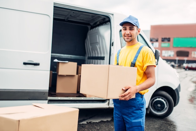 Workman or courier in uniform holds carton box in hands, truck with parcels