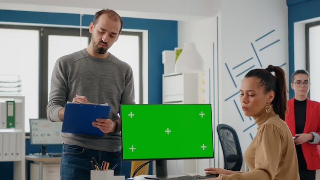 Workmates using computer with green screen on display in business office. Coworkers talking and working with isolated chroma key and blank mockup template on monitor. Copy space