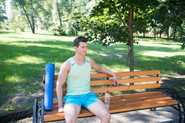 Workout is over. Man with yoga mat and water bottle sit on bench in park. Join outdoors yoga practice. Athlete with yoga equipment relaxing in park. Man cares about health, he chose yoga outdoors.