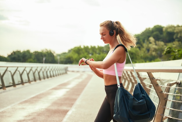 ワークアウトの結果、ヘッドホンでバッグを持ち、スポーツブレスレットを見ている若いスポーティな女性