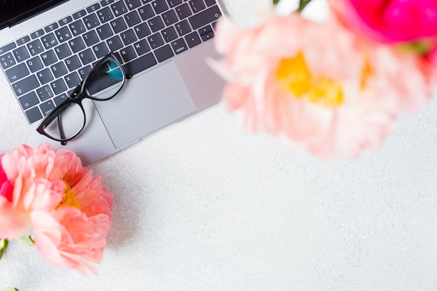 Workplace, desktop view. Laptop, pink peony and glasses on white