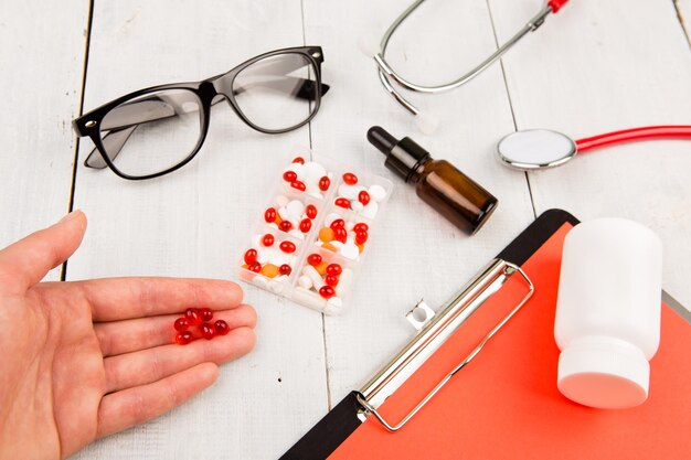 Workplace of a doctor Pills in hand red stethoscope clipboard and glasses on white wooden table