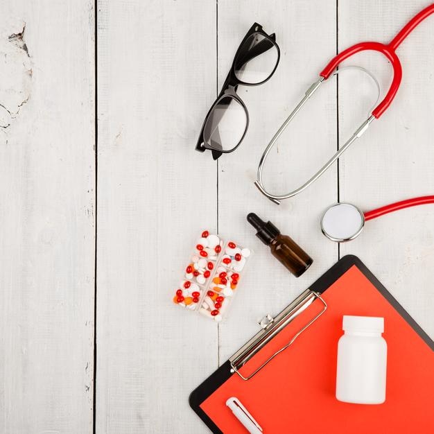 Workplace of a doctor Red stethoscope clipboard glasses and pills on white wooden table