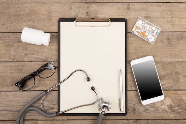 Workplace of a doctor Stethoscope clipboard pills smartphone and other stuff on wooden desk