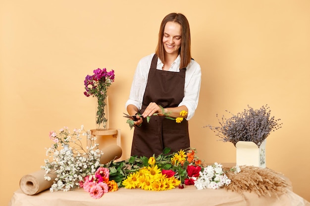 職場の花飾り 小売花屋 新鮮な花飾り 笑顔で満足した茶色のの女性 花屋が職場で茶色のエプロンを着て ベージュの壁にポーズをとっています
