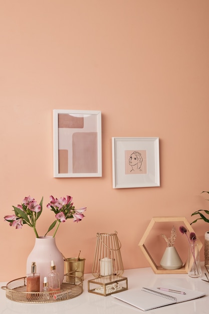 Workplace of interior designer by pink wall with two pictures in frames by table with flowers in vases, beauty care products, candles and other stuff