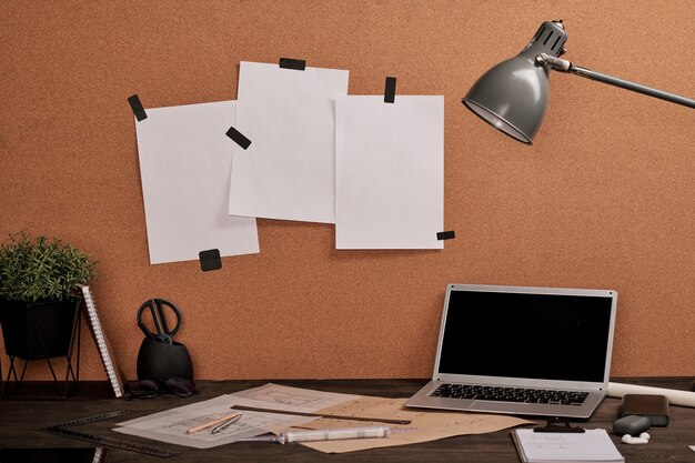 Workplace of modern engineer with blank papers on carton board in front of table with laptop, blueprints, lamp, rulers and other office supplies