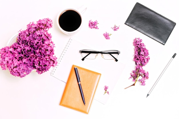 Workplace with cup of coffee, diary, pen, pencil, glasses and lilac flowers on the white table background. Flat lay, top view