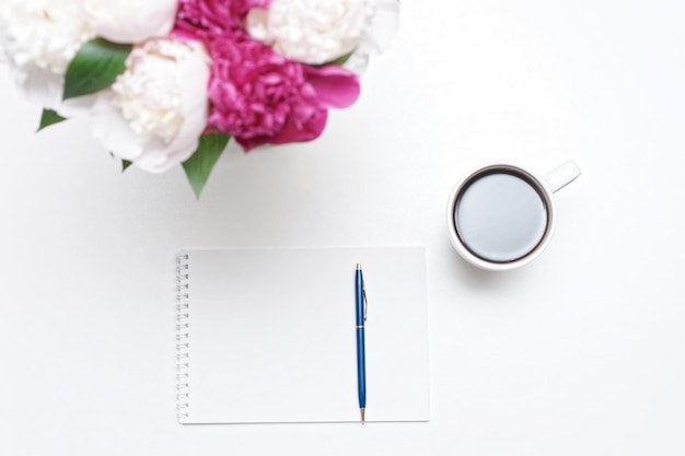 Photo workplace with cup of coffee, pen, paper and pink and white peony flowers on white table background. flat lay, top view