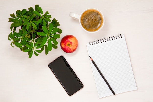 A workplace with a Notepad, phone, pencil, coffee Cup and potted flower is placed on a white background. Flat spoon, top view.