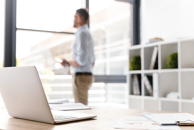 workplace with open white laptop lying on table, while defocused business man standing and looking through big window on