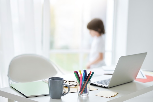 Workplace with white laptop, notes and cup of tea on the table at home. Bright light coming from the window. Little boy standing in the background. Interior design, home education concept
