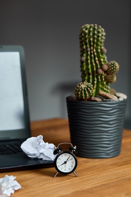 Workplace, wooden office desk with clock, sheet of paper, laptop, notebook, crumpled paper balls and supplies