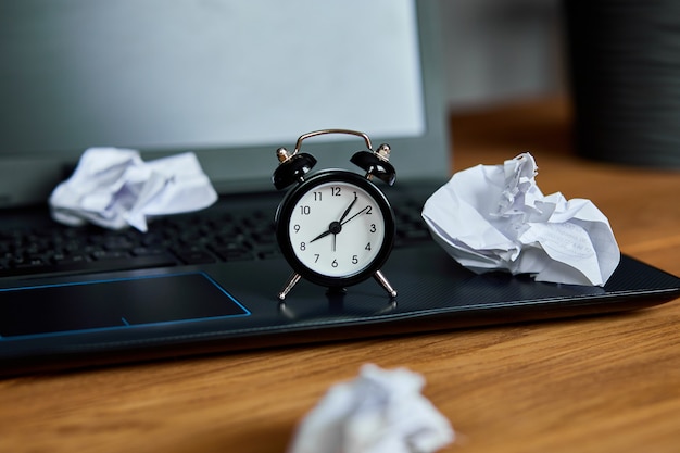 Workplace, wooden office desk with clock, sheet of paper, laptop, notebook, crumpled paper balls and supplies,